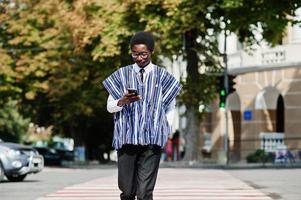 homme africain en vêtements traditionnels et lunettes marchant au passage pour piétons et regardant un téléphone portable. photo