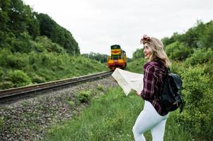 portrait d'une jeune blonde en chemise tartan à côté du train avec une carte. photo