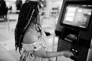 jolie fille afro-américaine de petite taille avec des dreadlocks, porter une robe jaune colorée, contre un guichet automatique avec carte de crédit à portée de main. noir et blanc. photo