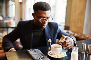 un homme afro-américain à la mode en costume et lunettes verse de la crème dans le café au café. photo