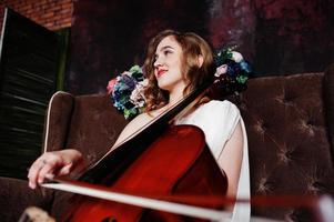 jolie jeune musicienne gilrl en robe blanche avec contrebasse assise sur un canapé marron vintage. photo