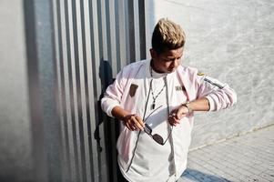 homme arabe hipster élégant à lunettes de soleil posé en plein air dans la rue au coucher du soleil contre un mur gris, regardant ses montres. photo