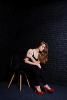 belle fille brune portant des talons hauts noirs et rouges, assis et posant sur une chaise au studio contre un mur de briques sombres. portrait de modèle de studio. photo