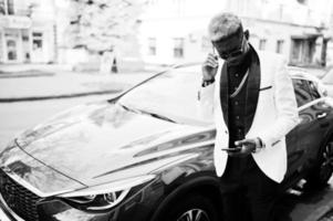 chic bel homme afro-américain en costume blanc contre une voiture de luxe rouge avec un téléphone portable à portée de main. photo