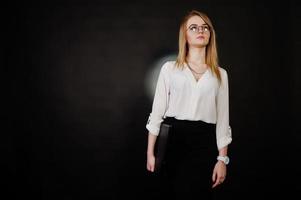 portrait en studio d'une femme d'affaires blonde à lunettes, chemisier blanc et jupe noire tenant un ordinateur portable sur fond sombre. femme réussie et concept de fille élégante. photo