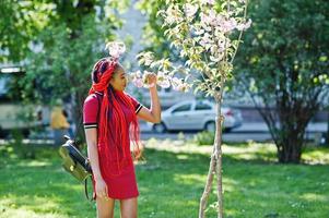 jolie et mince fille afro-américaine en robe rouge avec des dreadlocks posés en plein air dans le parc du printemps. modèle noir élégant. photo