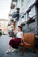 fille modèle blonde à la mode et belle en élégante jupe en velours rouge, chemisier blanc et chapeau, posée sur un banc dans la rue de la ville. photo