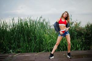 fille modèle bouclée sexy en haut rouge, short en jean jeans, veste en cuir et baskets posées dans le roseau. photo