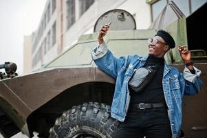 homme afro-américain en veste de jeans, béret et lunettes, fumant un cigare et posé contre un véhicule blindé militaire btr, faisant du selfie au téléphone. photo