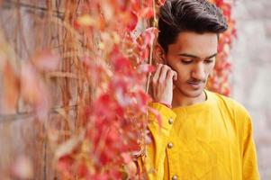 homme élégant indien en vêtements traditionnels jaunes posés en plein air contre le mur de feuilles rouges. photo