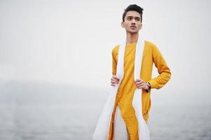 homme élégant indien en vêtements traditionnels jaunes avec foulard blanc posé en plein air contre la jetée brumeuse de la mer. photo