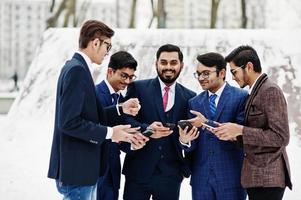 groupe de cinq hommes d'affaires indiens en costumes posés en plein air en journée d'hiver en europe, regardant sur les téléphones et disscus. photo