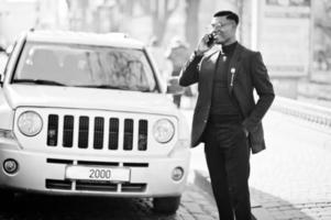 un homme afro-américain à la recherche étonnante porte un blazer bleu avec une broche, un col roulé noir et des lunettes posées dans la rue. gars noir à la mode parlant au téléphone contre la voiture. photo