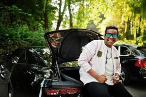 Un homme arabe hipster élégant à lunettes de soleil posé en plein air dans la rue et assis sur le coffre de sa voiture de muscle noire. homme noir riche. photo