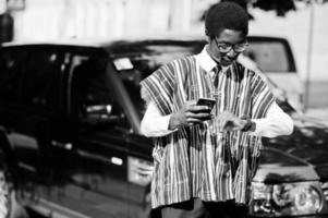 homme d'affaires africain en vêtements traditionnels et lunettes avec téléphone portable contre suv de voiture noire regardant sur ses montres. Africains riches. photo