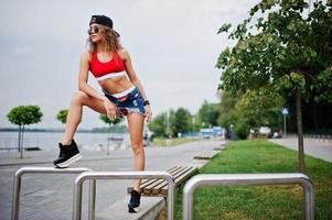 fille modèle bouclée sexy en haut rouge, short en jean, lunettes de soleil coupe et baskets posées contre des rails en acier. photo