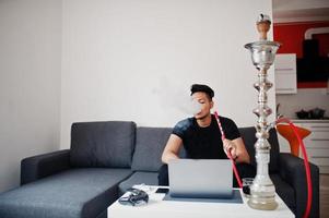 homme indien beau et à la mode en noir assis dans la chambre, fumant du narguilé et travaillant sur un ordinateur portable. photo