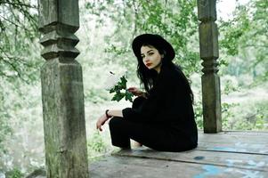 fille sensuelle tout en noir, lèvres rouges et chapeau. femme dramatique goth tenir fleur de chrysanthème blanc avec libellule. photo