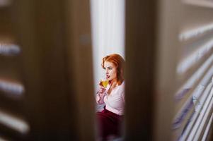 magnifique fille aux cheveux rouges en chemisier rose et jupe rouge avec un verre de vin à portée de main sur le lit de la chambre. photo