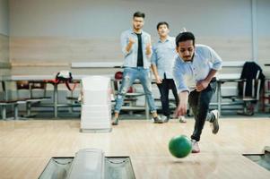 homme sud-asiatique en chemise jeans debout au bowling avec ballon sur les mains et le jeter. ses amis le soutiennent bruyamment. photo