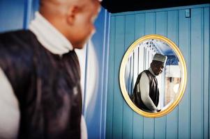 hommes africains en vêtements traditionnels noirs avec casquette regardant miroir sur mur bleu. photo