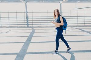 jolie jeune femme se promène dehors, porte un sac à dos, tient un téléphone portable moderne, écoute de la musique, vérifie la chanson préférée dans la liste de lecture, porte des vêtements de sport décontractés et des baskets, a un mode de vie sain photo