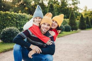 bel homme a de bonnes relations avec sa femme et sa petite fille qui l'embrassent de dos, ont des sourires agréables. une jeune famille sympathique en bonnets et chandails tricotés se tient contre des arbres verts photo