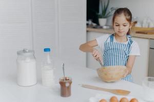 enfants, cuisine et concept de maison. jolie fille aux cheveux noirs fouette les ingrédients dans un bol, occupée à préparer la pâte pour le gâteau, étant le futur chef, porte un t-shirt blanc et un tablier rayé, utilise des ingrédients photo