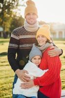 portrait vertical d'un couple familial amical se tenir ensemble, s'embrasser, avoir de bonnes relations, profiter du temps ensoleillé. bel homme mal rasé embrasse sa femme et sa fille, pose à l'extérieur photo