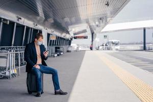 un homme européen est assis à la valise sur la station de quai de bus, essaie de trouver l'horaire de transport en ligne sur smartphone, attend le transport, voyage pendant une situation de pandémie dangereuse, porte un masque médical