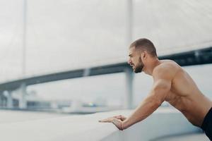 un sportif masculin motivé pousse des poses d'exercice en plein air au pont concentré dans la distance a un torse musclé nu d'entraînement du matin se réchauffe avant de faire du jogging. concept de mode de vie sain. photo