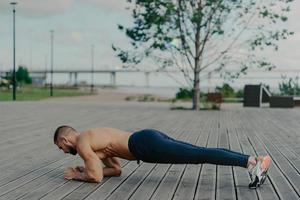 photo horizontale d'un homme barbu sportif se tient en pose de planche, pratique le yoga en plein air et vêtu de vêtements actifs, respire l'air frais. concept de sport, de fitness et de mode de vie sain. séance d'entraînement