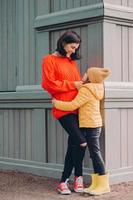 photo verticale d'une jolie mère attentionnée regarde les yeux de sa fille avec amour, embrasse tout en posant à l'extérieur, porte un jean déchiré, un pull et des baskets, sourit positivement. notion de relation familiale