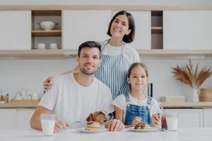 trois membres de la famille et leur animal de compagnie posent tous ensemble à la cuisine, mangent des crêpes sucrées au chocolat, boivent du lait, une femme au foyer affectueuse pose derrière le mari et la fille, embrasse, prépare le petit déjeuner photo