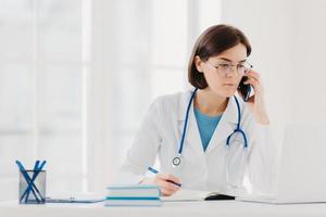 personnel de soins de santé, concept de médecine. femme médecin brune sérieuse concentrée sur un ordinateur portable moderne, réécrit les informations nécessaires, parle sur un téléphone portable, appelle quelqu'un, a un regard sérieux photo