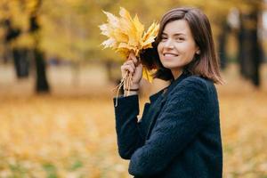 photo en plein air d'une femme assez détendue respire l'air frais, étant de bonne humeur, porte des feuilles jaunes, vêtue d'un manteau, pose contre l'espace de copie pour votre publicité. concept de saison et de journée ensoleillée