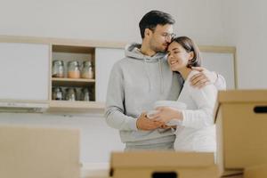 jeune couple amoureux s'embrasse et s'embrasse avec tendresse, tient une pile d'assiettes blanches, se tient dans la cuisine pendant le jour du déménagement, entouré de nombreuses boîtes en carton remplies d'effets personnels, déballe des trucs photo