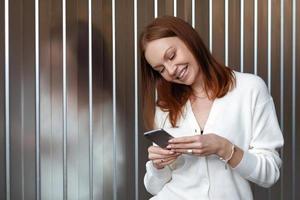 Une économiste européenne réussie et positive effectue un paiement en argent sur le site Web, pose avec un téléphone portable, vêtue d'une tenue blanche, sourit joyeusement, envoie un SMS, aime la communication en ligne. photo