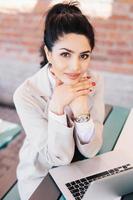 jeune femme d'affaires brune aux yeux charmants, mains douces avec manucure rouge portant une montre à portée de main et manteau blanc tenant les mains sous le menton assise près de son ordinateur portable se reposant après un travail acharné photo