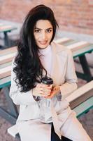 portrait d'une femme adorable avec de longs cheveux foncés ondulés, des yeux brillants et des lèvres pleines portant un manteau blanc assis sur un banc dans un café en plein air buvant du café à emporter posant à huis clos en souriant agréablement photo