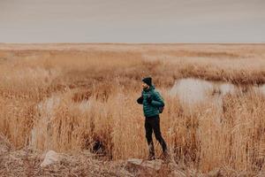 photo de côté d'un homme séduisant vêtu d'un chapeau, d'une veste et d'un pantalon à la mode, porte un sac à dos, se tient à l'extérieur, a un voyage inoubliable, profite de l'automne. concept de personnes, de repos et de style de vie