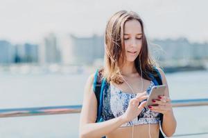 jolie fille active aux cheveux raides foncés, manucure rouge, porte un haut, porte un sac à dos, utilise le cellulaire moderne pour la messagerie et le divertissement, écoute la piste préférée dans les écouteurs, a un style de vie actif photo