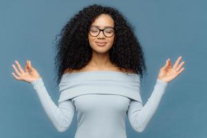 belle femme détendue avec une coiffure afro, se sent soulagée, montre un geste zen, garde les mains dans un geste correct, contrôle les émotions, prend le contrôle avec elle-même a les yeux fermés isolés sur fond bleu photo