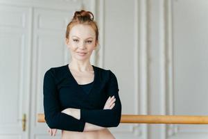 photo à la taille d'une danseuse de ballet satisfaite et confiante croise les mains sur la poitrine, porte un haut noir, a les cheveux roux, se tient près des barres, pose seule dans le centre de remise en forme, utilise une machine chorégraphique