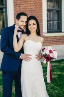 agréable jeune femme brune en robe de mariée blanche tient un bouquet près de son beau marié qui porte un costume formel, se tient à l'extérieur, pose à huis clos, a des expressions heureuses. photo