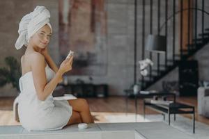 photo intérieure d'une belle femme tenant une bouteille de lotion a une peau lisse bien soignée porte une serviette sur la tête et autour du corps nu après avoir pris une douche, pose contre l'intérieur de la maison. notion de beauté.
