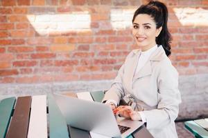 jolie étudiante brune portant un élégant manteau blanc travaillant à l'ordinateur écrivant sa thèse regardant avec le sourire à la caméra. femme d'affaires en vêtements élégants assis à l'intérieur sur un mur de briques photo