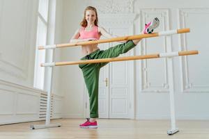 photo intérieure d'une danseuse rousse étire les jambes sur la barre dans un studio de ballet, porte un haut et un pantalon roses, des baskets roses, fait des exercices de fitness pilates, pose dans un studio de gym, prépare les muscles