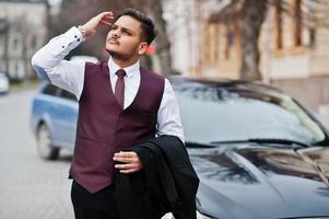 homme d'affaires indien élégant en costume de gilet de tenue de soirée debout contre une voiture d'affaires noire dans la rue de la ville. photo