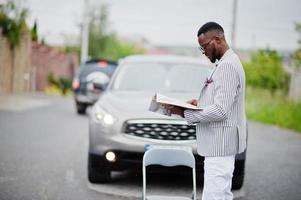homme afro-américain riche et élégant en blazer et pantalon blanc, lunettes lire le magazine contre sa voiture suv. photo